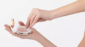 A hand holding a compact makeup case while another hand reaches inside to take some product. The compact has a sleek white design with a gold accent.