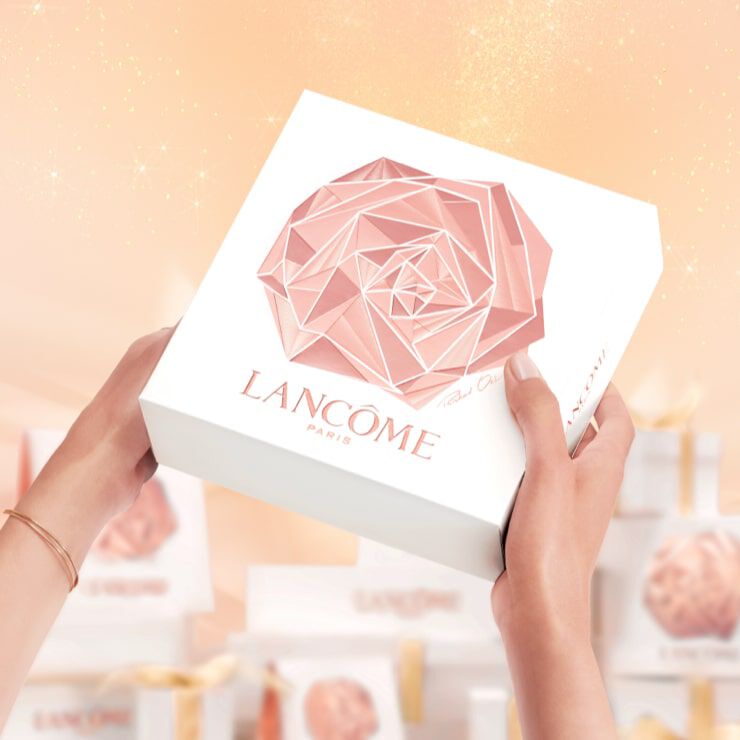 A person holding a white gift box featuring a geometric rose design and the Lancome Paris logo, with a soft, shimmering background.