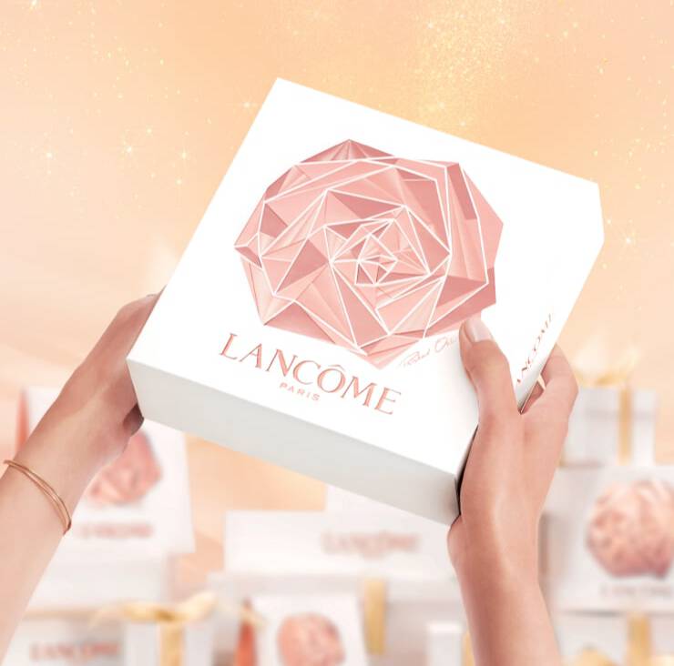 A person holding a white gift box featuring a geometric rose design and the Lancome Paris logo, with a soft, shimmering background.