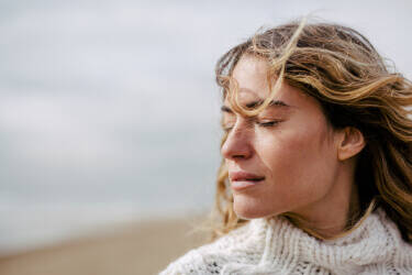 A woman with long, wavy hair stands outdoors with her eyes closed, looking serene against a blurred beach background. She is wearing a cozy, knitted white sweater.