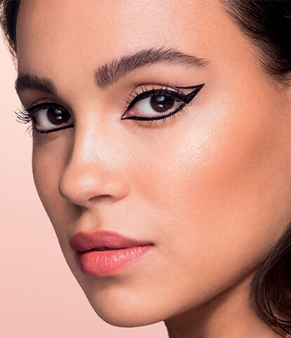A close-up profile of a woman with striking winged eyeliner and glowing skin, set against a soft pink background.