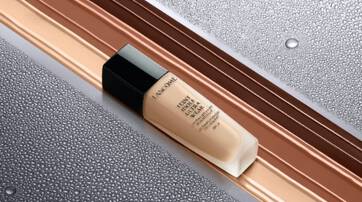 A bottle of foundation sits on a sleek, wet surface, with droplets of water highlighting the elegant packaging. The background features a gradient of warm tones.