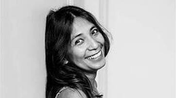A smiling woman with long, dark hair leaning against a wall, captured in black and white.