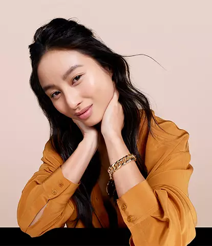 A young woman with long, dark hair resting her chin on her hands, smiling softly against a light beige backdrop. She is wearing a stylish brown shirt.