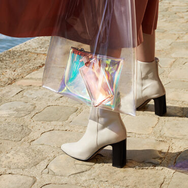 A close-up of a person's legs wearing white ankle boots, holding a transparent bag with an iridescent clutch inside, standing on a cobblestone surface near water.
