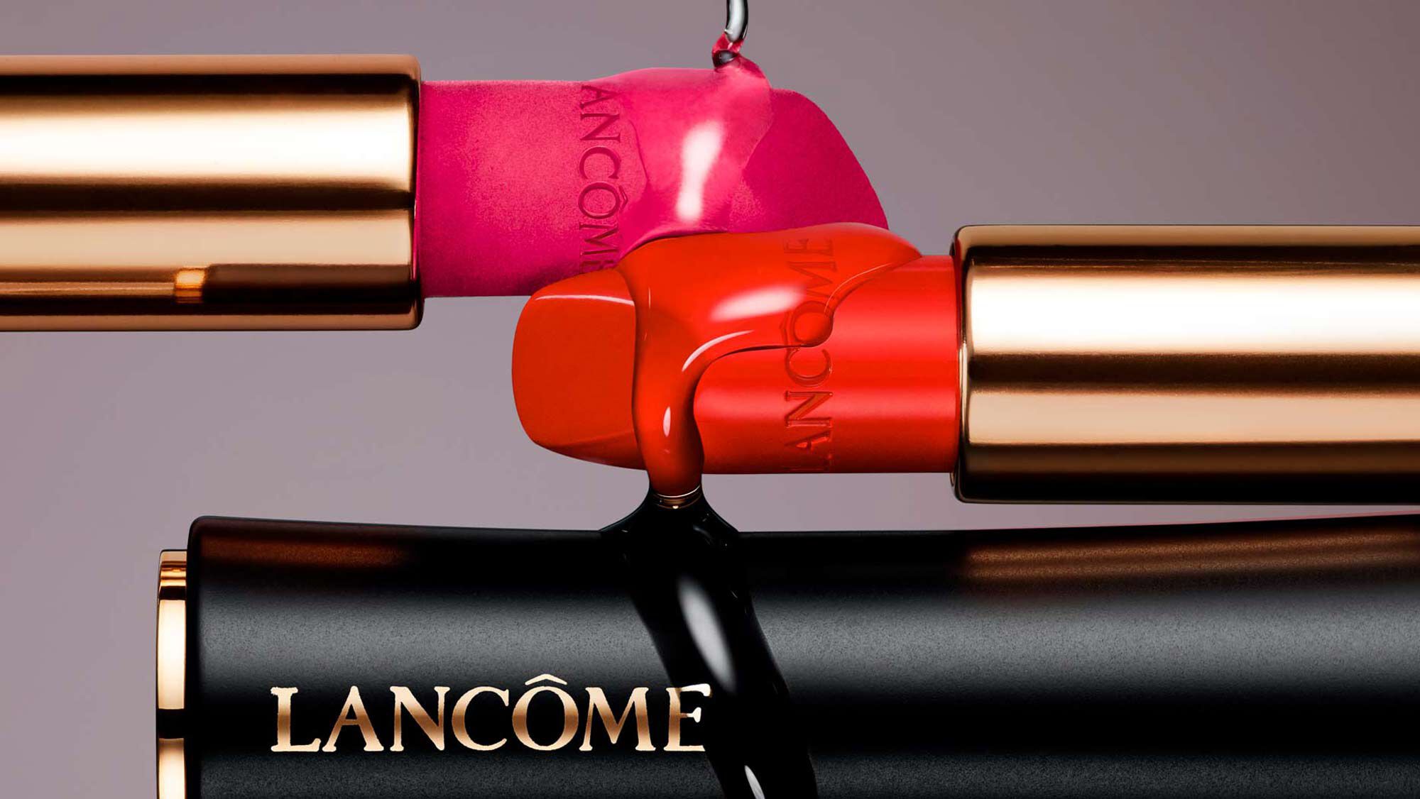 A close-up of three lipsticks in vibrant pink, red, and black cases, showcasing the Lancome logo. The lipsticks have unique, artistic shapes and are positioned to highlight their colors against a soft gray background.