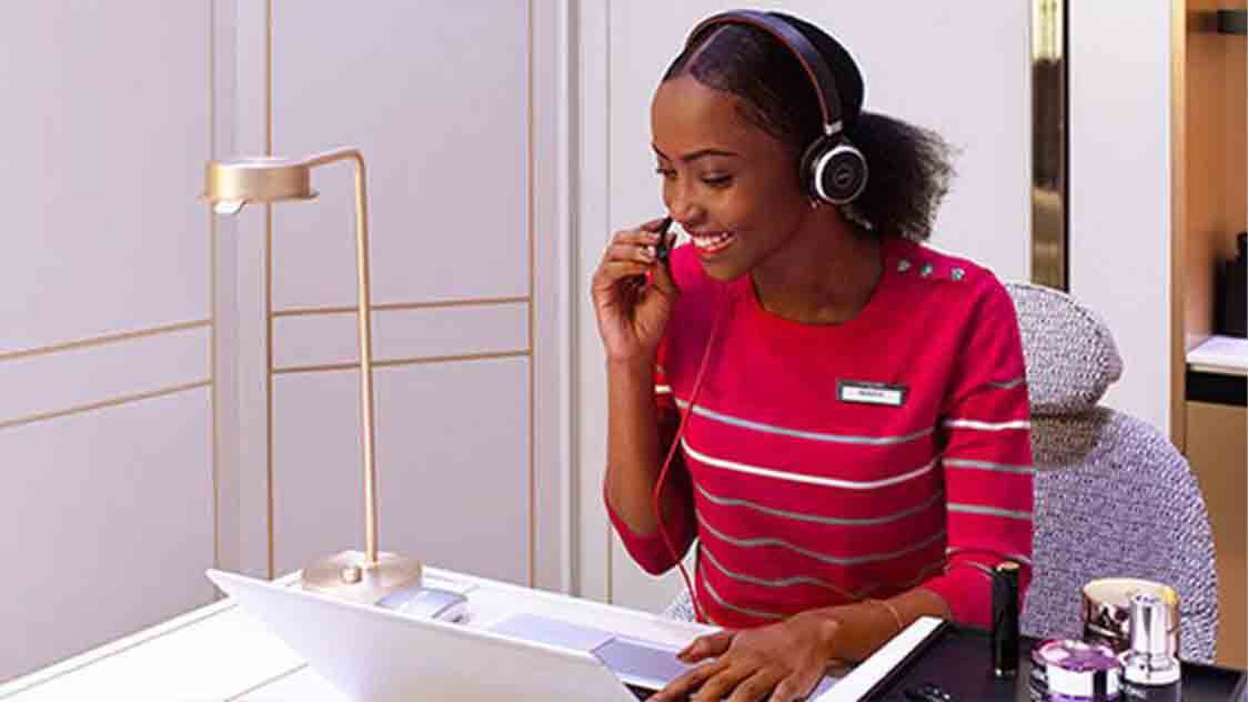 A woman wearing a red striped shirt and headphones smiles while speaking on a headset at a workstation with a laptop and cosmetics in the foreground.