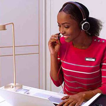 A woman wearing a red striped shirt and headphones smiles while speaking on a headset at a workstation with a laptop and cosmetics in the foreground.