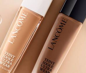 Two bottles of Lancome foundation are displayed against a soft beige background, with droplets of water nearby. The bottles are labeled "Teint Idole Ultra Wear" and "Care & Glow."