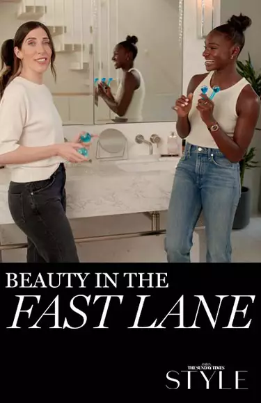 A smiling woman holding beauty products stands next to another woman who is engaging with her in a stylish bathroom setting. The text overlay reads "Beauty in the Fast Lane."