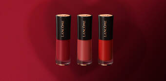 Three Lancome lipstick tubes displayed against a deep red background, showcasing shades of red.