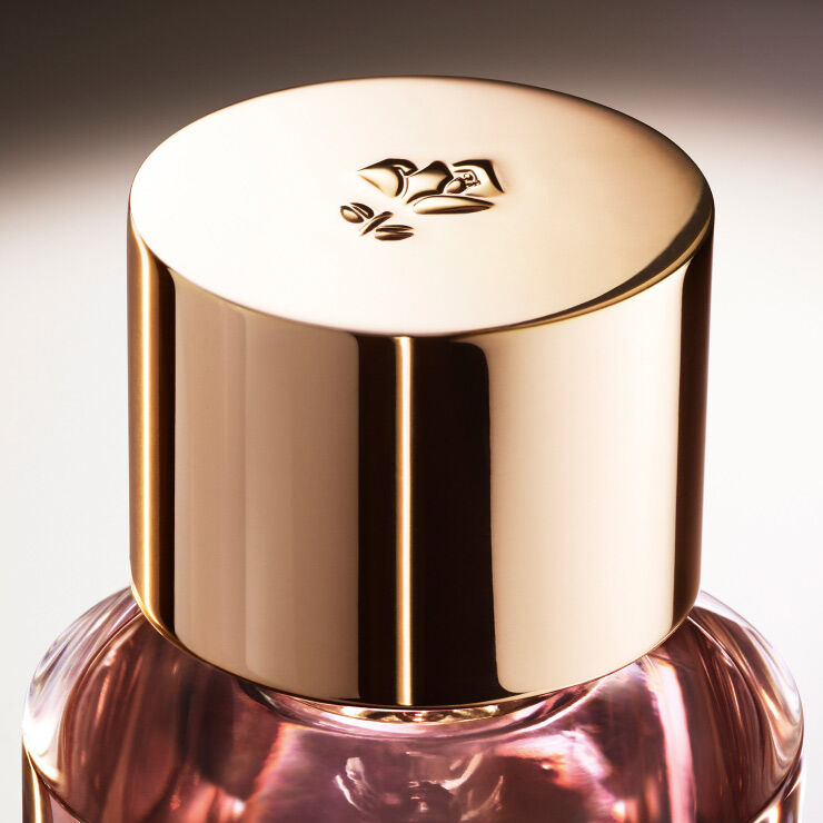 Close-up of a perfume bottle with a shiny gold cap, featuring a floral emblem on top, against a softly blurred background.