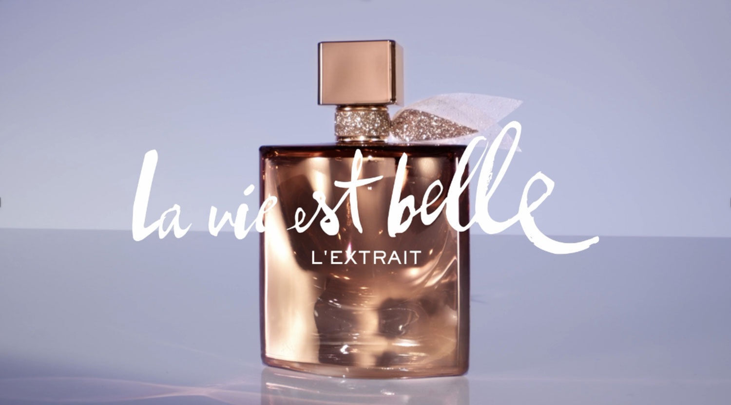 A elegant perfume bottle with a sophisticated design, featuring a geometric cap and a soft ribbon, set against a light backdrop. The text "la vie est belle l'extrait" is artistically displayed in a stylish font.