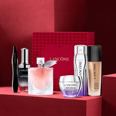 A collection of Lancome beauty products displayed on a textured red background, including a foundation, skincare jars, a perfume bottle, and a mascara.