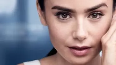 A close-up of a woman with dark hair and natural makeup, looking at the viewer with her hand near her face, against a blurred blue background.