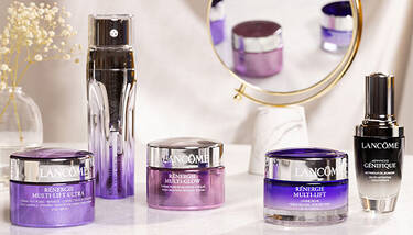 A collection of Lancome skincare products, including various Renergie and Advanced Genifique items, are elegantly arranged on a white surface with a small vase of baby's breath and a round mirror reflecting some of the products in the background.