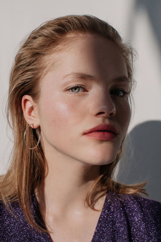 A close-up portrait of a young woman with straight blonde hair, wearing hoop earrings. She has a natural makeup look and is gazing directly at the camera, with soft lighting creating shadows on her face.