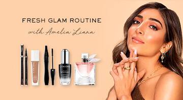 A woman with wavy hair and a glamorous makeup look poses confidently. She holds a lipstick near her mouth, surrounded by beauty products including a perfume, foundation, and eyeliners. The text "Fresh Glam Routine with Amelia Liana" is displayed on the left. The background is a soft peach color.