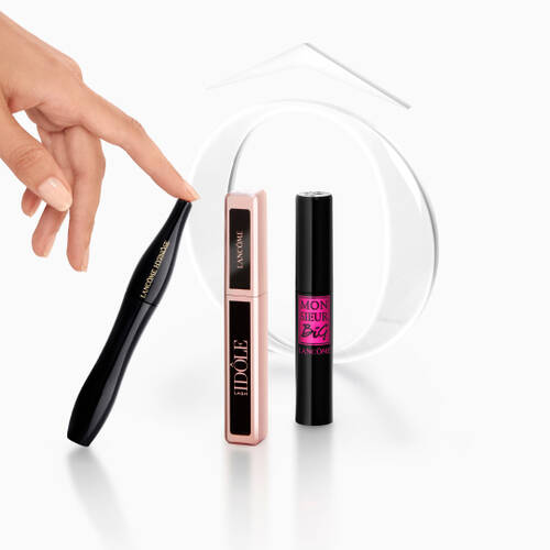A hand pointing at three different mascara products from Lancome arranged on a reflective surface, with a decorative clear object in the background.