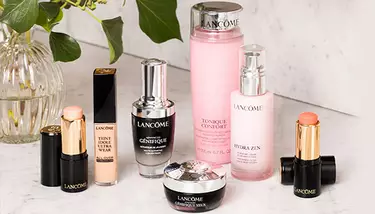 A variety of Lancome beauty products, including foundation sticks, serums, toners, and moisturizers, are arranged on a white marble surface next to a glass vase with greenery.