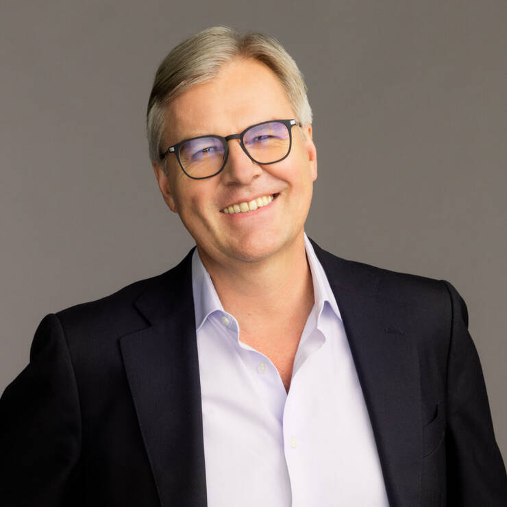 A smiling man with light gray hair and glasses, dressed in a black blazer and light shirt, against a neutral backdrop.