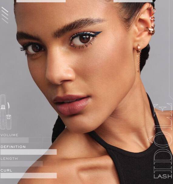 A close-up portrait of a woman with a strong jawline and braided hair, showcasing bold blue eyeliner and a natural lip color. She wears a stylish black top and gold earrings, with minimalist graphics highlighting makeup attributes around her.