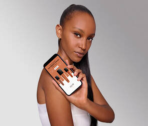 A person with long black hair holds a smartphone displaying different shades of foundation against a gradient background.