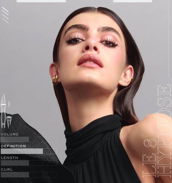 A close-up portrait of a confident woman with long, sleek hair and bold makeup, wearing a high-neck black top. The image features a minimalist design with labels for volume, definition, and length alongside the product name "LE 8 HYPNOSE."