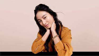A young woman with long dark hair, wearing an orange-brown top and a gold bracelet, rests her chin on her hands and smiles gently, against a beige background.