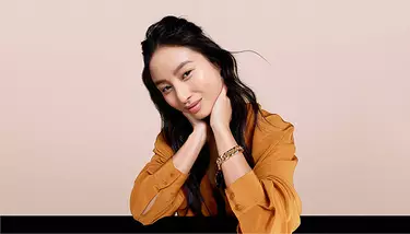 A young woman with long dark hair, wearing an orange-brown top and a gold bracelet, rests her chin on her hands and smiles gently, against a beige background.