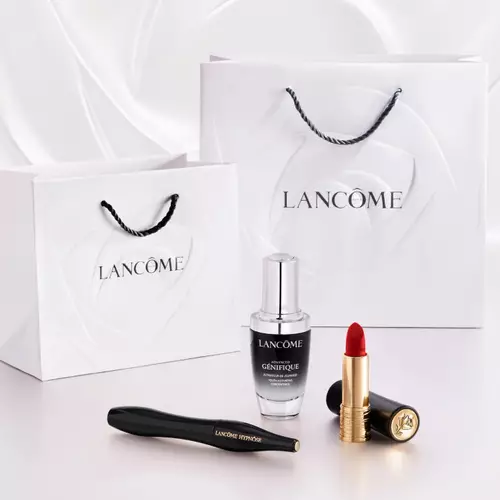 A white gift bag with the Lancome logo, accompanied by a bottle of serum, a tube of red lipstick, and a pair of tweezers on a light background.