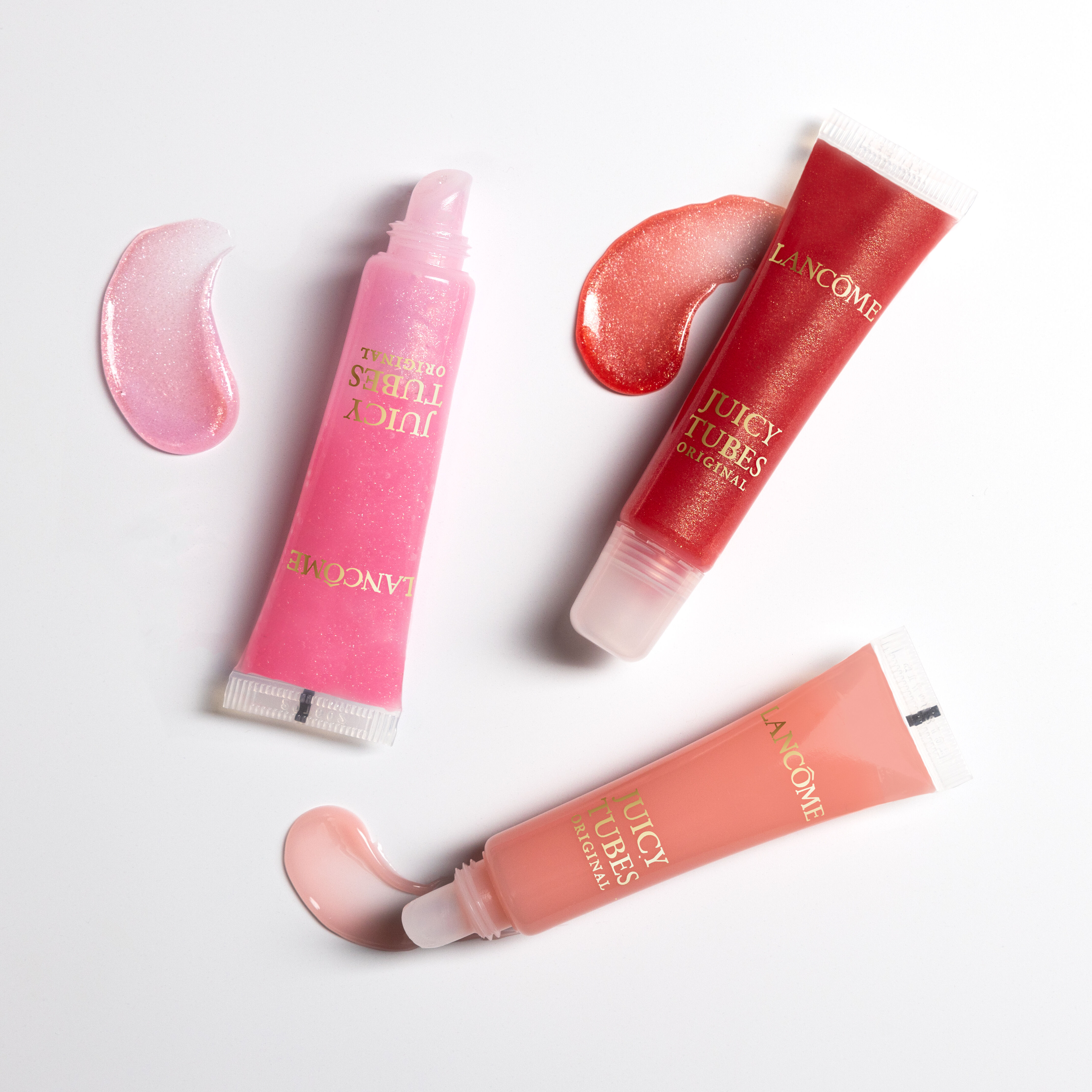 Three tubes of Lancome Juicy Tubes lip gloss in pink, red, and peach shades, with corresponding swirls of gloss beside them.