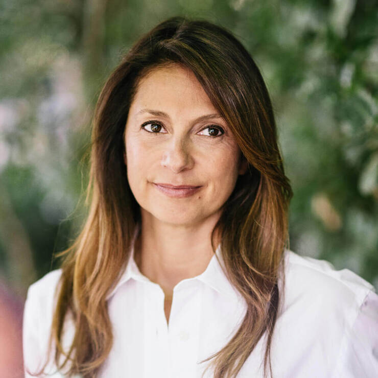 A close-up portrait of a woman with long, styled hair, wearing a white shirt, against a blurred green background.