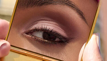 A close-up of an eye with subtle pink and brown eyeshadow, dark eyeliner, and mascara, reflected in a gold compact mirror held by manicured fingers.