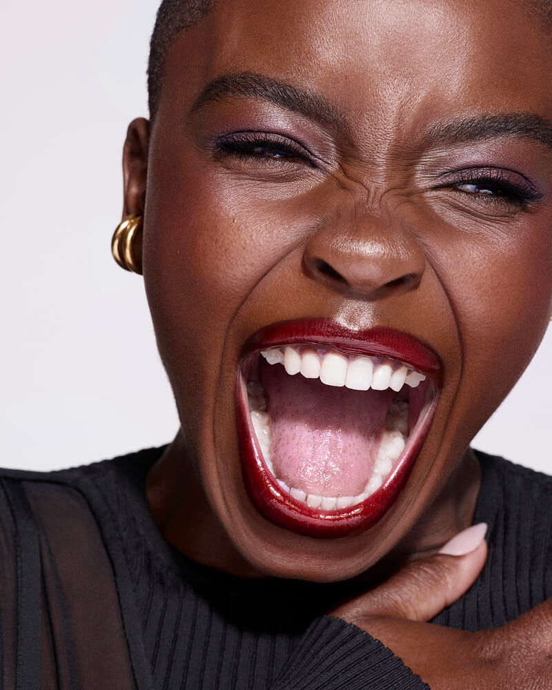 A close-up of a person with short hair, wearing bold red lipstick, smiling widely and laughing, showcasing expressive eyes and ear jewelry against a plain background.