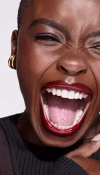 A close-up of a person with short hair, wearing bold red lipstick, smiling widely and laughing, showcasing expressive eyes and ear jewelry against a plain background.