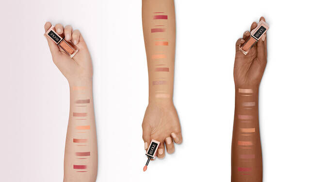 Three arms displaying swatches of various lip colors on different skin tones, with one hand holding a lip product.