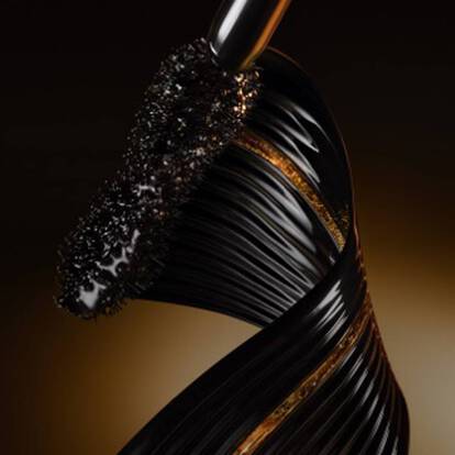 An artistic close-up of a glossy black mascara brush against a gradient golden background, highlighting its sleek design and shimmering texture.
