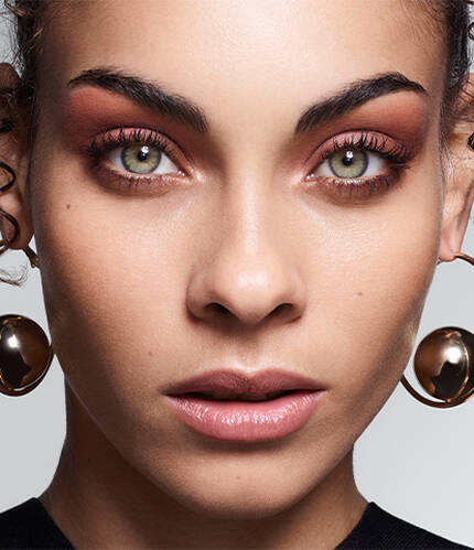 A close-up portrait of a person with curly hair and striking green eyes, wearing large gold hoop earrings and a dark top, against a softly lit gray background.