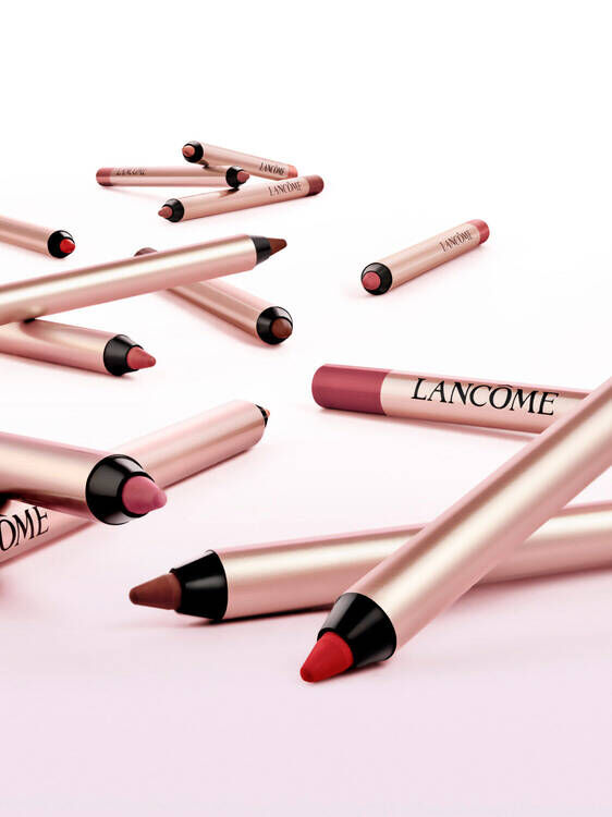 A collection of Lancome lip crayons in various shades, scattered on a soft pink background. The crayons feature a sleek design with the Lancome logo prominently displayed.