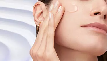 A woman with clear skin and a gold hoop earring gently touches her cheek, where a drop of translucent product is visible, against a soft, light background.