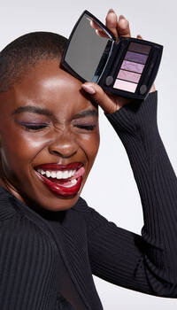 A smiling woman with short hair holds an eyeshadow palette up to her face, showcasing vibrant eye colors. She is wearing a black ribbed top and has bold red lipstick on.