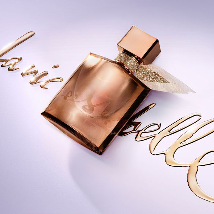 A close-up of a perfume bottle with a sleek, rose gold design, placed next to elegant script spelling "la vie est belle" in a stylish font. The background is a soft lavender color.