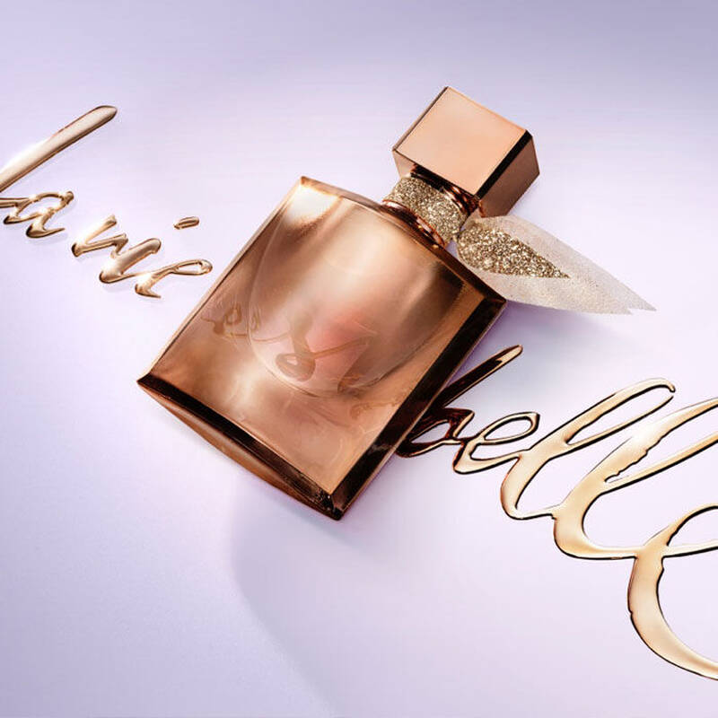 A close-up of a perfume bottle with a sleek, rose gold design, placed next to elegant script spelling "la vie est belle" in a stylish font. The background is a soft lavender color.