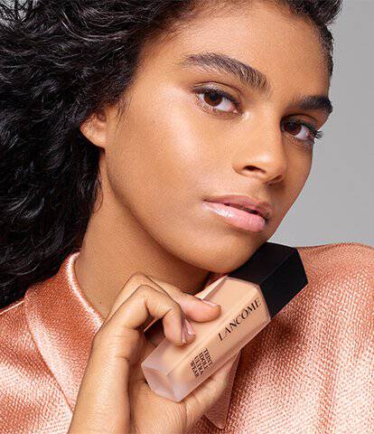 A model with curly hair poses confidently, holding a foundation bottle while wearing a shimmering, copper-toned top against a neutral background.