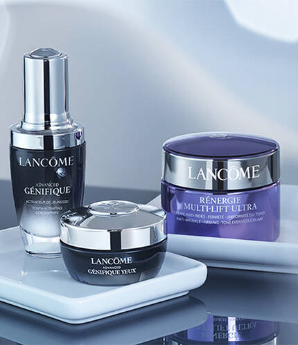 Three Lancome skincare products displayed on a clean, white surface, with a glass of water in the background. The products include a serum, a cream, and a gel cream, all elegantly packaged.