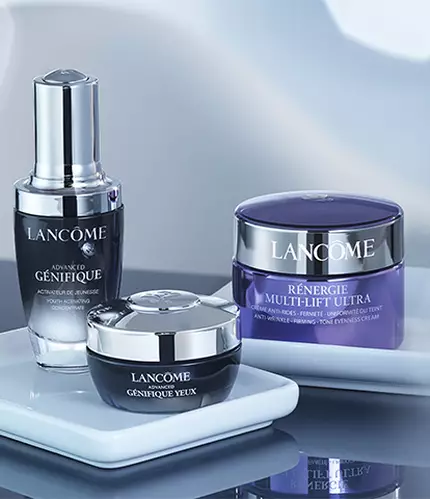 Three Lancome skincare products displayed on a clean, white surface, with a glass of water in the background. The products include a serum, a cream, and a gel cream, all elegantly packaged.