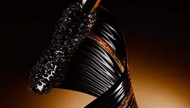 A close-up shot of a mascara wand coated in black product, alongside a sleek, twisted black and gold element, set against a dark, warm-toned background.