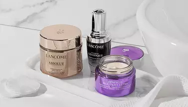 Lancome Absolue Soft Cream, Advanced Genifique serum, and Renergie Multi-Lift Ultra cream are displayed on a white tray with cotton pads, set against a marble background with a white sink.