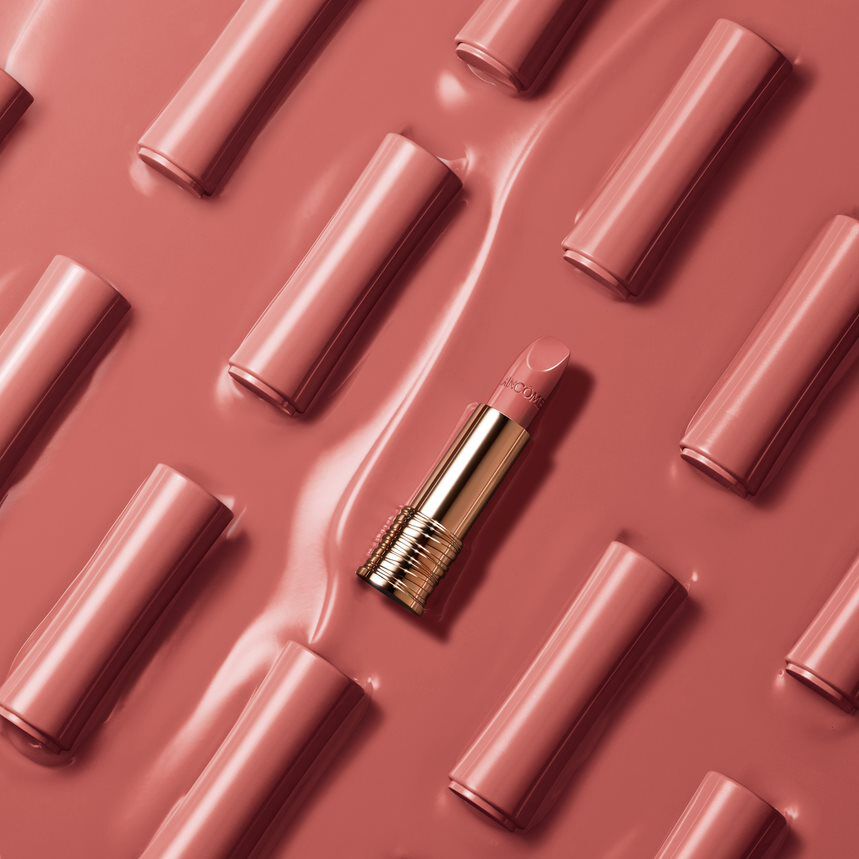 A close-up of a nude pink lipstick tube lying on a glossy pink background, surrounded by similar lipstick tubes arranged neatly.